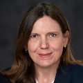 Portrait of person with dark hair smiling in a blue suit against a grey backdrop.