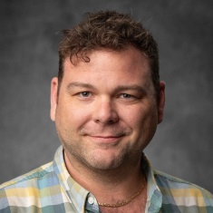 A person with short curly hair, smiling, wearing a plaid button-up shirt.