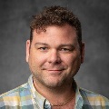 A person with short curly hair, smiling, wearing a plaid button-up shirt.
