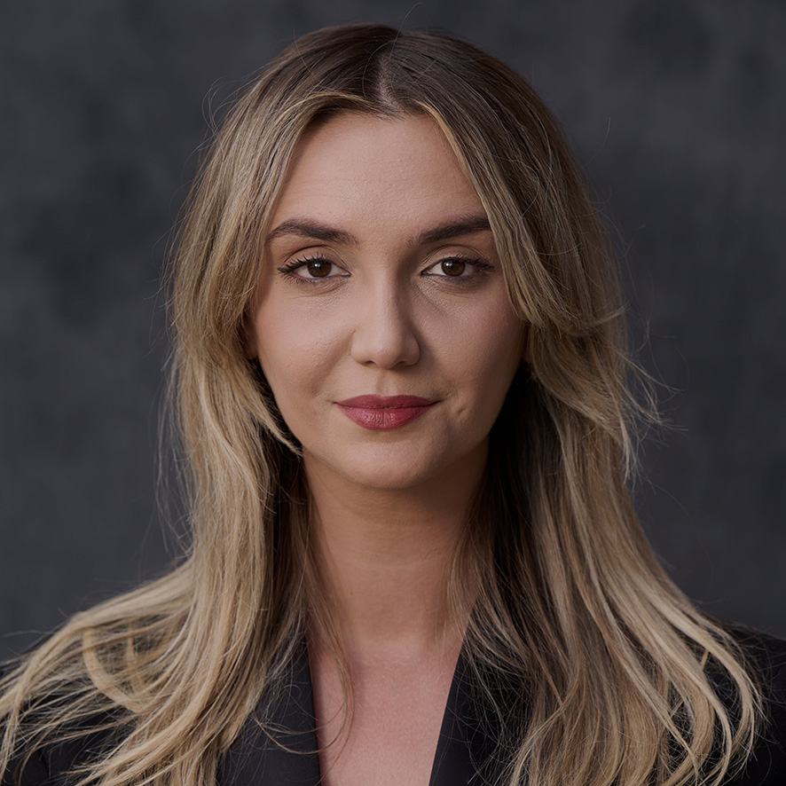 Portrait of a person with shoulder-length blond hair, wearing a dark blazer. They have a subtle smile and are looking directly at the camera against a dark gray background.