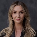 Portrait of a person with shoulder-length blond hair, wearing a dark blazer. They have a subtle smile and are looking directly at the camera against a dark gray background.