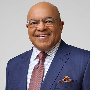 A person smiling. They are wearing glasses and are dressed in a navy blue suit jacket with a light plaid dress shirt, a mauve tie, and a patterned pocket square. The background is a neutral light gray.