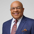 A person smiling. They are wearing glasses and are dressed in a navy blue suit jacket with a light plaid dress shirt, a mauve tie, and a patterned pocket square. The background is a neutral light gray.
