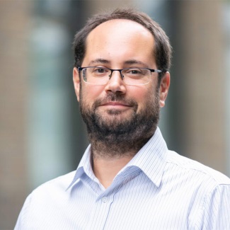 Person with glasses and a beard, wearing a blue shirt, smiling and standing in front of a blurred city background.