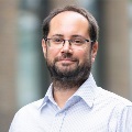Person with glasses and a beard, wearing a blue shirt, smiling and standing in front of a blurred city background.