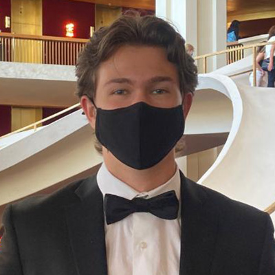 Person wearing a black mask and formal attire with a bow tie, posing in front of a staircase.