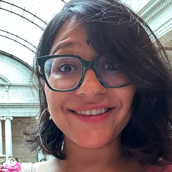 Close-up portrait of a smiling person wearing glasses, inside a building with an arched glass ceiling.
