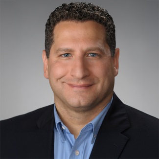 Professional headshot of a person with curly hair, wearing a dark blazer and a blue shirt, smiling at the camera.