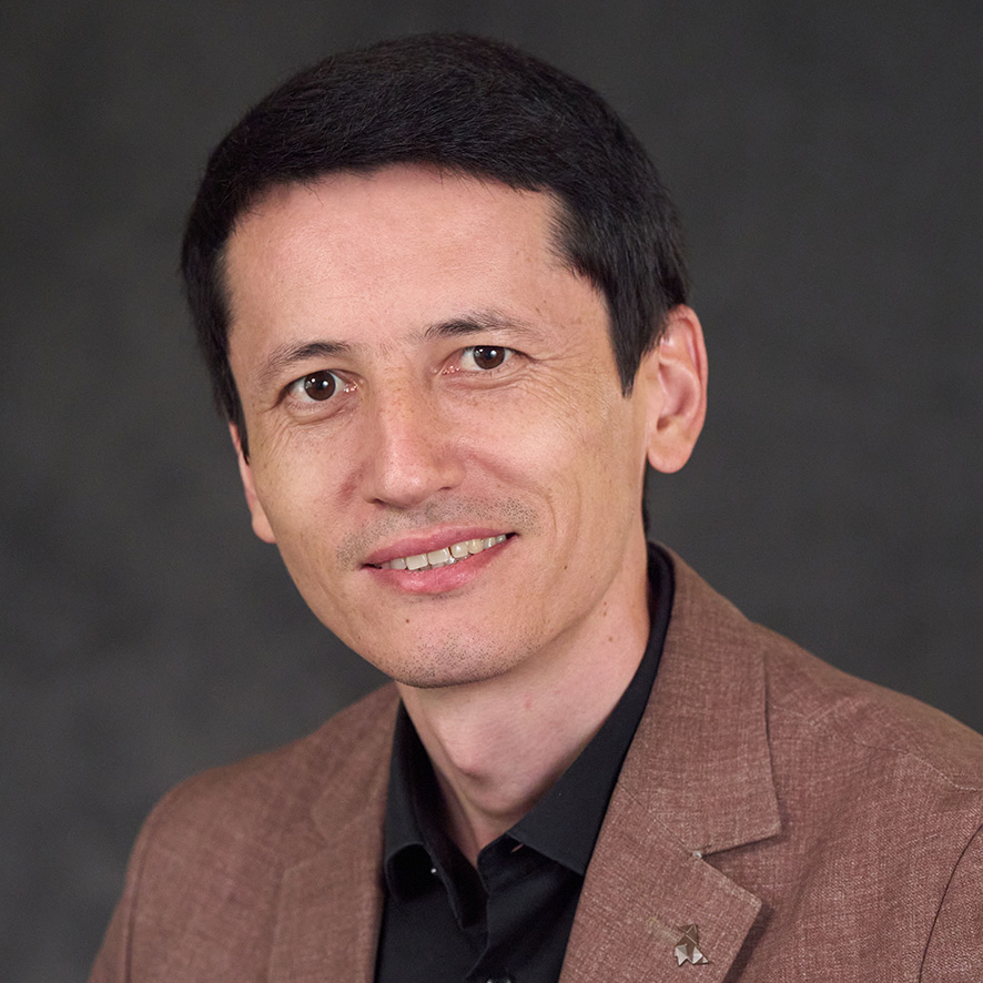 Portrait of a smiling person wearing a brown blazer and black shirt, featuring a pin with a star design.