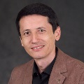 Portrait of a smiling person wearing a brown blazer and black shirt, featuring a pin with a star design.