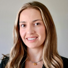 A person with long, wavy light brown hair with blonde highlights, smiling with a closed-mouth expression. They are wearing a dark blazer and a delicate necklace with a small pendant, photographed against a plain light gray background.