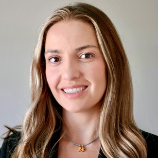 A person with long, wavy light brown hair with blonde highlights, smiling with a closed-mouth expression. They are wearing a dark blazer and a delicate necklace with a small pendant, photographed against a plain light gray background.