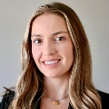 A person with long, wavy light brown hair with blonde highlights, smiling with a closed-mouth expression. They are wearing a dark blazer and a delicate necklace with a small pendant, photographed against a plain light gray background.