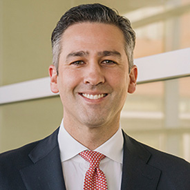 A professional portrait of a smiling individual wearing a suit and a red tie.