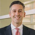 A professional portrait of a smiling individual wearing a suit and a red tie.