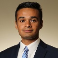 A person wearing a navy suit, white dress shirt, and a blue and white striped tie, set against a neutral beige background