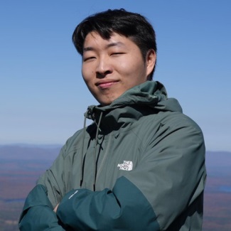 A person standing outdoors on a mountain summit, arms crossed, wearing a green jacket.