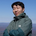 A person standing outdoors on a mountain summit, arms crossed, wearing a green jacket.