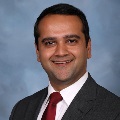 A person smiling and wearinga gray suit, white dress shirt and a red tie, set against a soft blue-gray background.