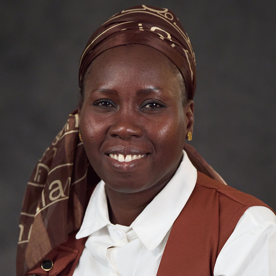 Portrait of a smiling individual wearing a brown headscarf and a white shirt under a brown vest.