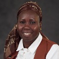 Portrait of a smiling individual wearing a brown headscarf and a white shirt under a brown vest.