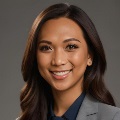 A person with long brown hair wearing gray blazer and navy blouse, smiling at camera against neutral background.