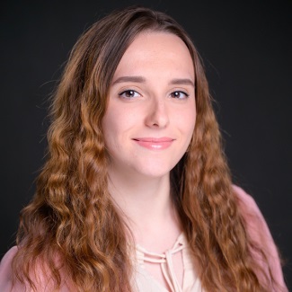 Professional portrait of a person with brown eyes and long reddish brown hair, wearing a pink top.