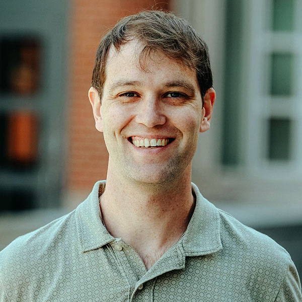 headshot photo of man standing outside, wearing a green shirt and smiling