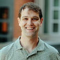 headshot photo of man standing outside, wearing a green shirt and smiling