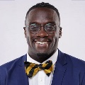 A person smiles broadly at the camera, wearing a navy blue blazer, white dress shirt, and black-and-gold striped bow tie. They have short hair and wears glasses, and is photographed against a light gray background.