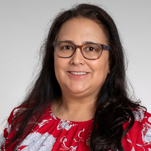 Portrait of a smiling individual with shoulder-length curly hair, wearing glasses and a red floral shirt.