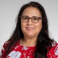 Portrait of a smiling individual with shoulder-length curly hair, wearing glasses and a red floral shirt.