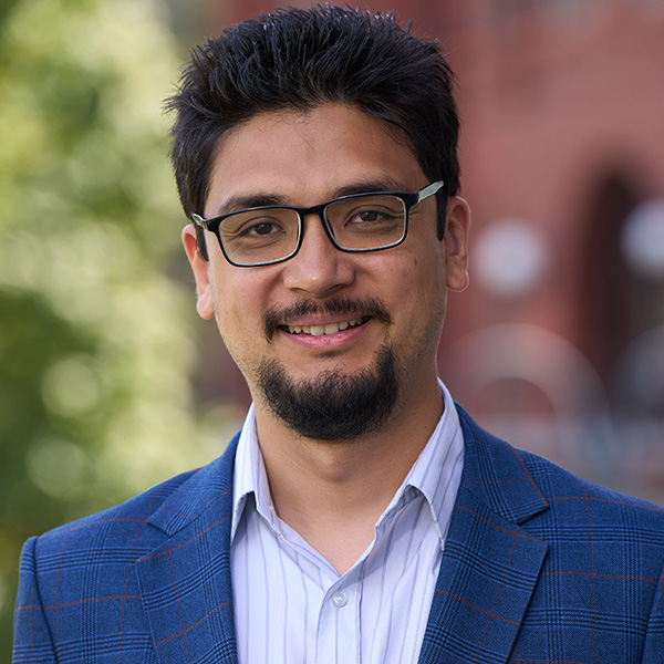 Portrait of a smiling person wearing glasses and a blue blazer, standing outdoors.