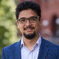 Portrait of a smiling person wearing glasses and a blue blazer, standing outdoors.