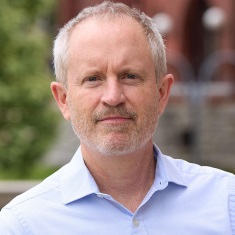 Portrait of a person smiling outdoors with a brick building in the background.