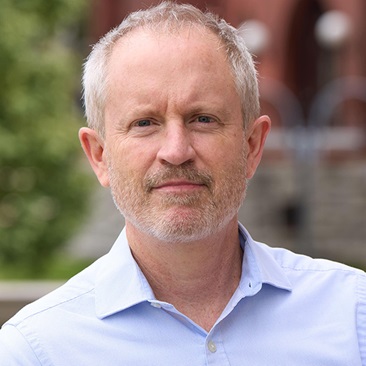 Portrait of a person smiling outdoors with a brick building in the background.