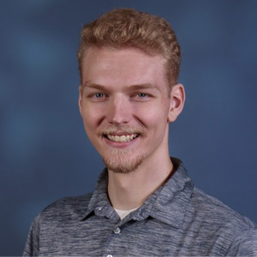 A person with curly reddish-blond hair and a light beard and mustache, smiling. They are wearing a heather gray polo shirt and standing against a dark blue textured background.