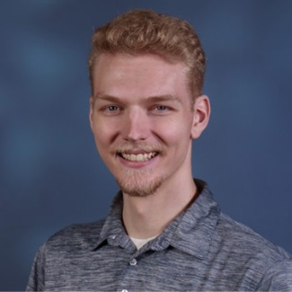 A person with curly reddish-blond hair and a light beard and mustache, smiling. They are wearing a heather gray polo shirt and standing against a dark blue textured background.