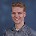 A person with curly reddish-blond hair and a light beard and mustache, smiling. They are wearing a heather gray polo shirt and standing against a dark blue textured background.