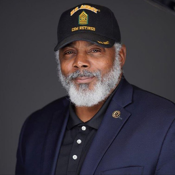 A person with a white mustache and beard wearing dress uniform and Command Sergeant Major Retired cap with gold embroidered insignia.
