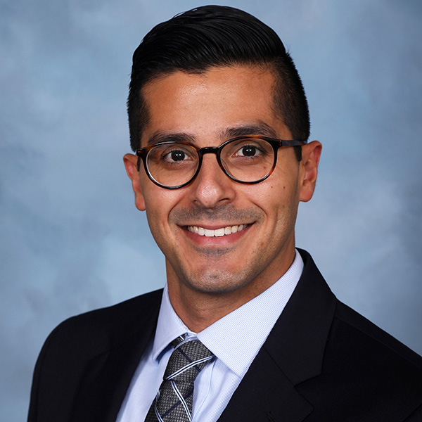 A person smiling and wearing dark-framed glasses, a black blazer, a light blue dress shirt and a gray patterned tie, set against a soft blue-gray background.