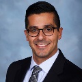 A person smiling and wearing dark-framed glasses, a black blazer, a light blue dress shirt and a gray patterned tie, set against a soft blue-gray background.