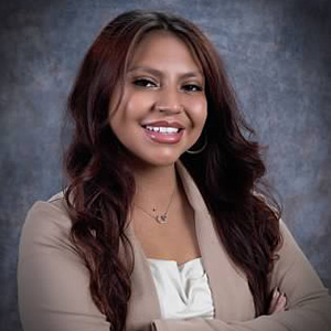 Professional portrait of a smiling person wearing a beige blazer and white top, set against a mottled grey background.