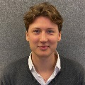 Portrait of a person with shoulder-length brown hair, wearing a white shirt and grey sweater.
