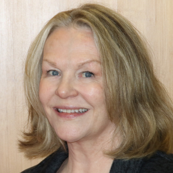 Portrait of a smiling person with shoulder-length blonde hair, standing in front of a wooden background.