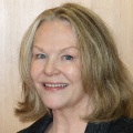 Portrait of a smiling person with shoulder-length blonde hair, standing in front of a wooden background.