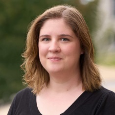 Headshot of an individual with shoulder-length brown hair smiling against a blurred outdoor background.