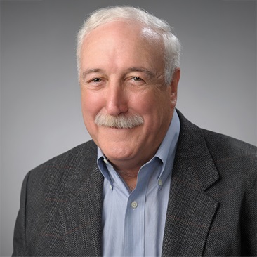 A person with short white hair and a white mustache, smiling, wearing a light blue button-down shirt and dark herringbone-patterned blazer, against a gray gradient background.