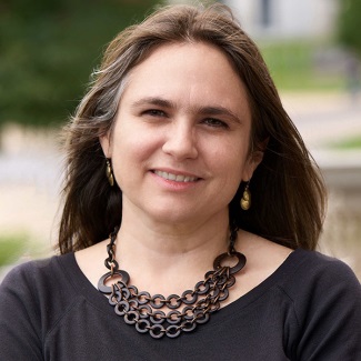 Portrait of a person smiling, wearing a dark top and a large chain necklace, with a blurred natural background.