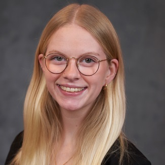 Portrait of a smiling individual with glasses, wearing a black outfit and gold earrings against a grey backdrop.